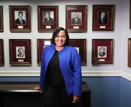 A woman in a blue suit jacket and black pants and shirt smiles at the camera while leaning on a desk in front of a wall of portraits