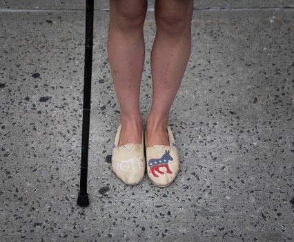 A photo of Rebecca from the knees down. She stands with her cane, wearing slip-on shoes with a democratic donkey on the left shoe and Vote written on the right shoe.