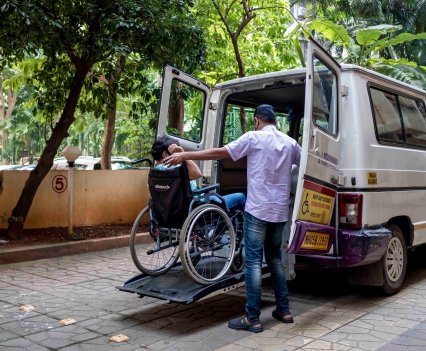 A man uses a lift to help a person in a wheelchair into a taxi van.