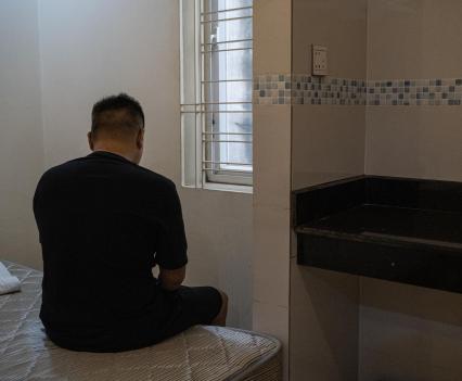 Man in a hunched over position sitting on the edge of a bed in a sparsely decorated room