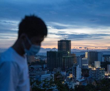 Young man wearing a mask, standing on a high point overlooking a city at sunset