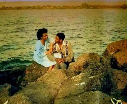 A man and woman sit on rocks by a large body of water