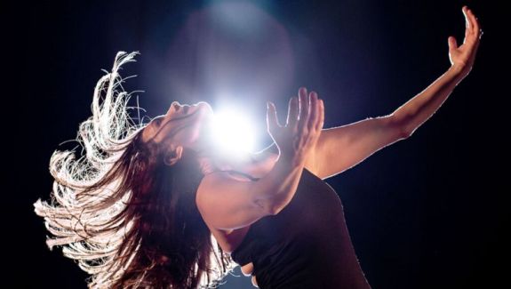 A woman with outstretched arms leans back with her hair flying out from her head. There is a bright light shining right above her.