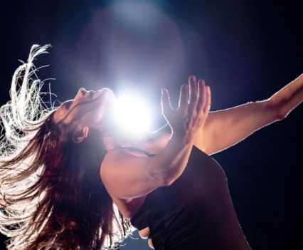 A woman with outstretched arms leans back with her hair flying out from her head. There is a bright light shining right above her.