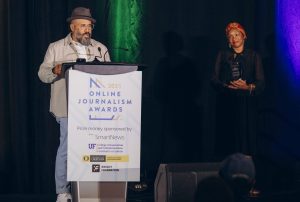 Robert Hernandez giving his speech behind a podium. Imaeyen Ibanga is to his right looking at him and smiling.