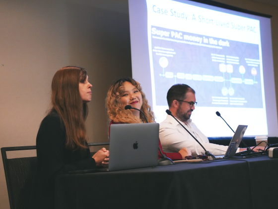 Three people sit in front of a presentation screen in a conference room. They are giving a presentation about open source intelligence.