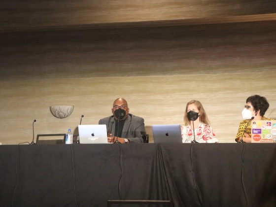 Three panelists sit at a black table with their computers.