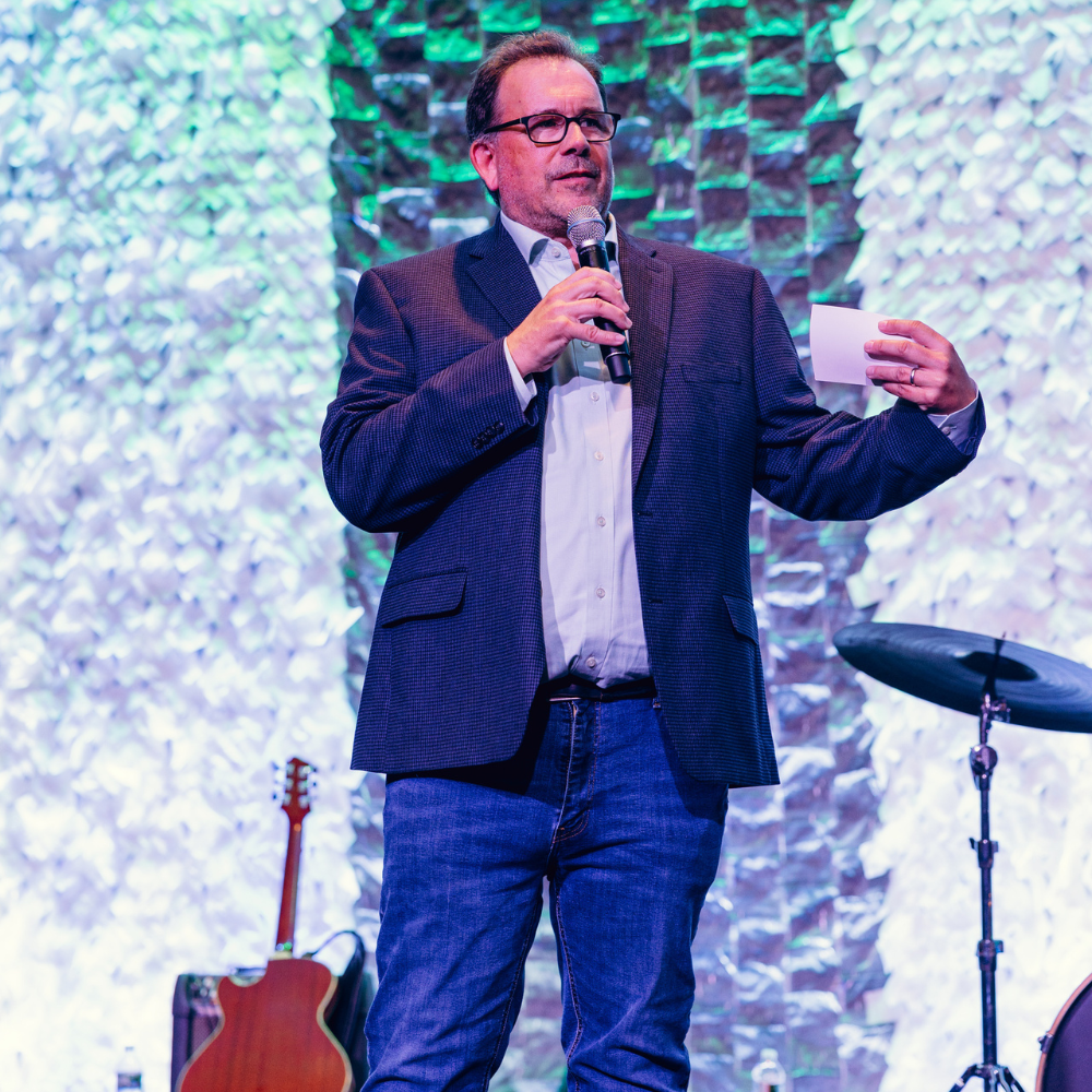 Jim Brady delivering remarks into a hand-held microphone on a stage.